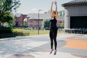 photo pleine longueur d'une jeune femme active saute haut garde les bras levés respire profondément porte un t-shirt noir des leggings et des chaussures de sport pose à l'extérieur sur le stade vous motive à faire du sport