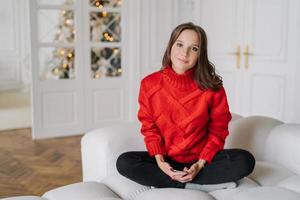 image d'une femme assez détendue vêtue d'un pull rouge décontracté, assise les jambes croisées sur un canapé blanc, tenant un téléphone intelligent, pose dans le salon avec un arbre de nouvel an décoré en arrière-plan, porte un pull rouge photo