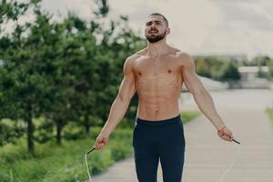 un homme athlétique motivé avec un torse nu concentré sur un programme d'entraînement physique intense saute par-dessus la corde à sauter, pose dans le parc a un corps sportif, respire l'air profondément. concept d'entraînement du matin photo