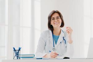 une jeune femme médecin écrit sur papier dans une clinique, crée une ordonnance pour le patient, fait de la paperasse à l'hôpital, porte une robe blanche médicale, est assise dans un bureau moderne et confortable. personnes, profession, concept de travail photo