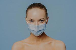 jeune modèle féminin sérieux regarde directement les supports de caméra épaules nues à l'intérieur porte un masque médical pour protéger la propagation du coronavirus isolé sur fond bleu. concept de soins de santé et de maladie photo