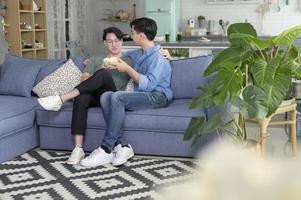 jeune couple gay souriant ayant du pop-corn en regardant la télévision dans le salon à la maison, lgbtq et concept de diversité. photo