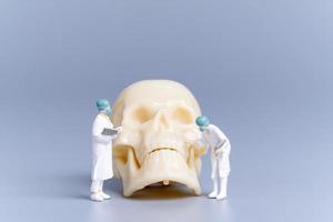 médecin de personnes miniatures avec un crâne humain géant sur fond gris photo