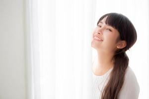 portrait d'une belle jeune femme asiatique debout à la fenêtre et souriant tout en se réveillant avec le lever du soleil le matin, fille heureuse avec fraîcheur et gaieté, mode de vie et concept de détente. photo