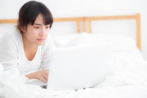 beau portrait asiatique jeune femme sourire allongé sur le lit à l'aide d'un ordinateur portable avec heureux dans la chambre pour les loisirs et se détendre, indépendant avec un cahier de travail fille, concept de communication. photo