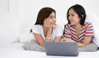 les jeunes couples de lesbiennes lgbt asiatiques utilisent des ordinateurs portables pour rechercher des informations afin d'acheter une nouvelle maison. tout en s'embrassant au lit. photo