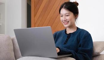 fille asiatique travaillant sur un ordinateur portable en ligne, utilisant internet, assise sur un canapé à la maison, espace libre photo