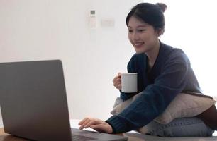 portrait de sourire heureux joyeux belle jolie femme asiatique se détendre boire et utiliser un ordinateur portable. fille abattant profiter de prendre le petit déjeuner en vacances matin vacances sur canapé à la maison photo