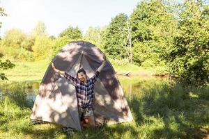 une femme heureuse dans une chemise à carreaux regarde par une tente touristique lors d'une randonnée sur la rive du fleuve le matin. camping en pleine nature, nuitée en pleine nature, vacances en famille et aventures. photo