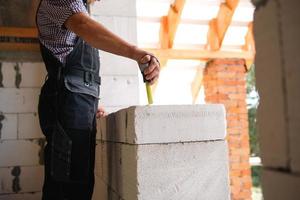 un ouvrier du bâtiment sur le chantier mesure la longueur de l'ouverture de la fenêtre et du mur de briques avec un ruban à mesurer. chalet sont faits de blocs de béton poreux, vêtements de travail - combinaison et casquette de baseball