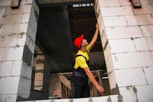 un ouvrier du bâtiment sur le chantier mesure la longueur de l'ouverture de la fenêtre et du mur de briques avec un ruban à mesurer. chalet sont faits de blocs de béton poreux, vêtements de travail - combinaison et casquette de baseball photo