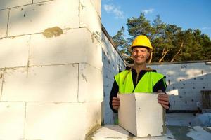 le constructeur tient entre ses mains un bloc de béton cellulaire - la maçonnerie des murs de la maison. travailleurs de la construction portant des vêtements de protection - un casque et un gilet réfléchissant. photo