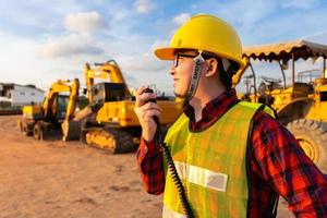 technicien civil ingénieur asiatique parle avec l'équipe par talkie-walkie et pointe au point d'enquête d'inspection pour le projet de travaux de construction de transport avec pelle sur fond de site d'ingénierie photo