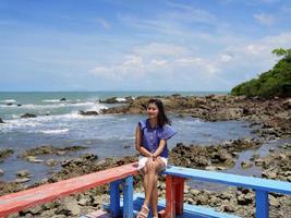 cheveux venteux femme femme touriste s'asseoir sur un siège en bois rouge et bleu coin terrasse avec vue sur la mer plage de roche et ciel bleu vif photo