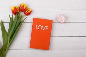 l'amour du livre rouge se trouve sur un tableau blanc. fleurs tulipes et cadeau avec un arc sur la table. photo