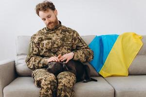 un soldat ukrainien en uniforme militaire est assis sur un canapé avec son fidèle ami, un chien amstaff, sur le fond du drapeau jaune et bleu. photo