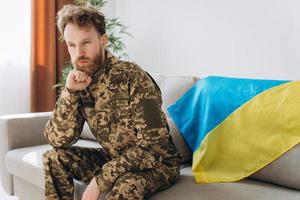 portrait d'un jeune soldat patriote ukrainien émotif en uniforme militaire assis sur le bureau sur un canapé avec un drapeau jaune et bleu. photo