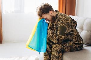 portrait d'un jeune soldat patriote ukrainien émotionnel en uniforme militaire assis sur le bureau sur le canapé tenant un drapeau jaune et bleu. photo