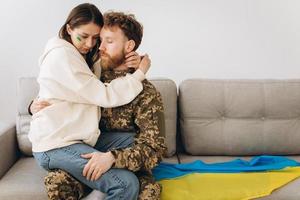 couple ukrainien, militaire en uniforme avec sa petite amie sur le canapé à la maison sur fond de drapeau jaune et bleu photo