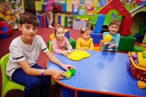 quatre enfants jouant dans un centre de jeux intérieur. jardin d'enfants ou salle de jeux préscolaire. assis près de la table avec des fruits en plastique. photo