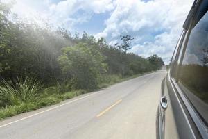 à côté de la voiture sur la route goudronnée. voiture roulant sur une route goudronnée avec de l'herbe verte et des arbres densément. voyage de voyage à la nature. sous le ciel bleu et les nuages blancs. photo