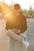 un jeune homme élégant dans un sweat à capuche, un pantalon décontracté et des baskets blanches. un gars avec un casque est assis et utilise un smartphone. photo aux tons orange.