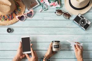 jeune couple tenant un téléphone intelligent à écran vide avec accessoires et articles d'été de voyage sur fond en bois avec espace de copie, concept de planification de voyage photo