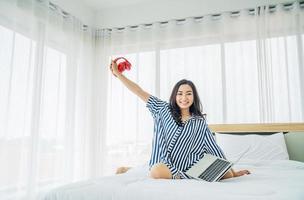 femme asiatique assise dans un lit confortable sous une couverture blanche. très heureuse avec ses bras levés et tenant des écouteurs après avoir écouté de la musique et travaillé sur son ordinateur portable, concept technologique et travail à la maison. photo