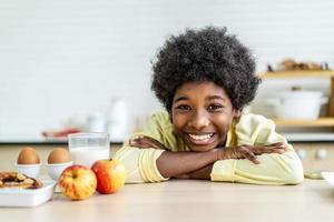 gros plan souriant petit garçon boire un verre de lait, assis à une table en bois dans la cuisine, adorable enfant enfant appréciant le yaourt biologique, obtenir des vitamines et du calcium, concept de soins de santé pour enfants photo