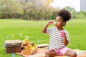 heureuse petite fille afro-américaine aux cheveux bouclés soufflant des bulles de savon jouant seule dans le parc, style de cheveux noirs photo