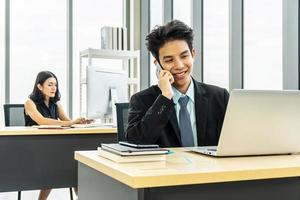 jeune homme d'affaires assis au bureau parlant sur un téléphone portable, assis au bureau, utilisant un ordinateur portable, souriant, femme d'affaires travaillant sur un ordinateur portable dans son poste de travail en arrière-plan. photo