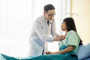 médecin de sexe masculin asiatique portant un stéthoscope sur l'oreille et prenant le rythme cardiaque d'une patiente assise sur un lit d'hôpital photo