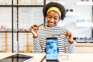 jeune femme afro-américaine entrepreneur est mode vlogger montrer des lunettes au téléphone portable et souriant le vendre en ligne en direct dans la boutique de lunettes photo