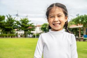 portrait d'une jolie petite fille asiatique debout dans un parc d'été regardant à huis clos souriant joyeusement, enfant riant, expressions faciales expressives. photo