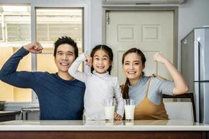 famille heureuse s'amusant dans la cuisine. père asiatique, mère et petite fille passent du temps ensemble et prennent leur petit déjeuner en buvant et en tenant des verres de lait à table photo