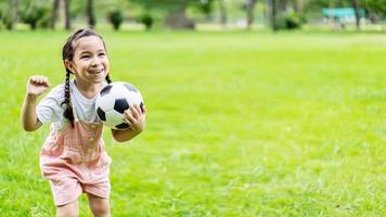 petite fille souriante debout tenant le ballon de football sur le terrain de football vert en été. portrait d'athlète de petite fille jouant avec un ballon au stade. concept d'enfance active. copie espace photo