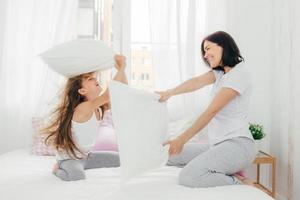 photo intérieure d'une femme européenne à l'air agréable a de la joie avec sa petite fille, a une bataille d'oreillers dans la chambre, pose dans une pièce lumineuse spacieuse et confortable, étant de bonne humeur. concept de personnes et d'amusement