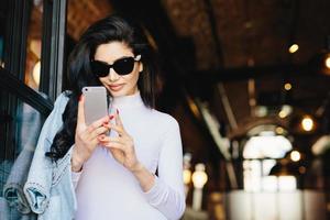 femme brune élégante à la peau pure, portant des lunettes de soleil, ayant une manucure parfaite rouge portant des vêtements formels blancs tenant un téléphone portable utilisant Internet gratuit, vérifiant ses e-mails. personnes, communications photo