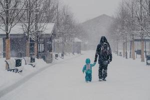 photo extérieure d'un petit enfant et d'un père couvrant la distance, étant sur le chemin du retour à la maison, se tenant la main, profitant du temps enneigé de l'hiver. vue arrière des personnes marchant dans la rue pendant les chutes de neige. notion de saison froide