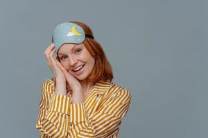 matin et se réveiller. jolie rousse joyeuse jeune femme européenne porte un bandeau et des vêtements de nuit, regarde avec plaisir la caméra, isolée sur un mur de studio gris avec un espace vide, se réveille de bonne humeur. photo