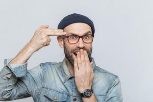 drôle d'homme barbu imbécile à l'intérieur, fait semblant de s'embrasser avec une arme à feu ou de faire un geste de suicide, couvre la bouche avec la main, regarde joyeusement dans la caméra, isolé sur fond gris. fous masculins photo