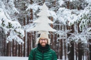 plan horizontal d'un modèle masculin souriant tenant un sapin sur la tête, se tenant à l'extérieur dans la forêt d'hiver, profitant du calme et de l'atmosphère tranquille. homme mal rasé de race blanche avec une expression joyeuse photo