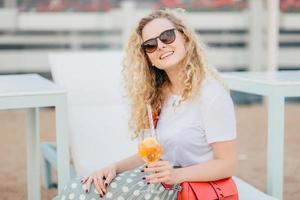 jolie jeune femme aux cheveux ondulés, porte des lunettes de soleil, se repose bien pendant l'été, tient une boisson fraîche, a un sourire amical, regarde positivement la caméra. personnes, boisson, concept de loisirs photo