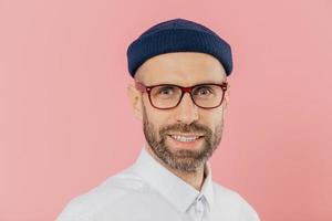 gros plan d'un homme mal rasé à l'air agréable porte des lunettes, a un sourire doux, regarde avec une expression faciale satisfaite à la caméra, porte une chemise blanche, isolé sur fond rose. designer masculin photo