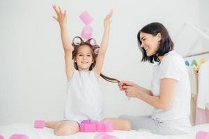 joyeuse jolie fille avec des bigoudis sur la tête, lève les mains, passe du temps libre avec sa mère, qui fait de la coiffure, étant à la maison. belle mère et fille s'amusent à l'intérieur, se coiffent. notion de beauté photo