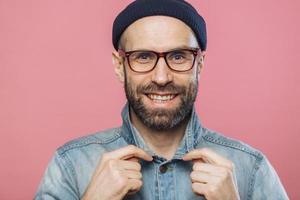 photo horizontale d'un homme barbu souriant et agréable portant des lunettes et une veste en jean, annonce de nouveaux vêtements à la mode, a une expression heureuse, isolé sur fond rose. Émotions positives