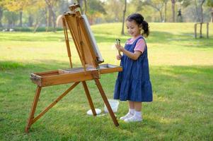 une petite fille est debout sur l'herbe et peinte sur la toile posée sur un pupitre à dessin photo