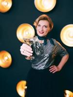 femme élégante et élégante mûre avec un verre de vin mousseux sur fond sombre de lampes. fête, célébration, concept anti-âge photo