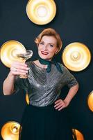 femme élégante et élégante mûre avec un verre de vin mousseux sur fond sombre de lampes. fête, célébration, concept anti-âge photo