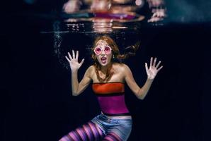 femme drôle rousse dans des vêtements colorés et des lunettes de soleil roses sur le fond sombre nageant sous l'eau photo